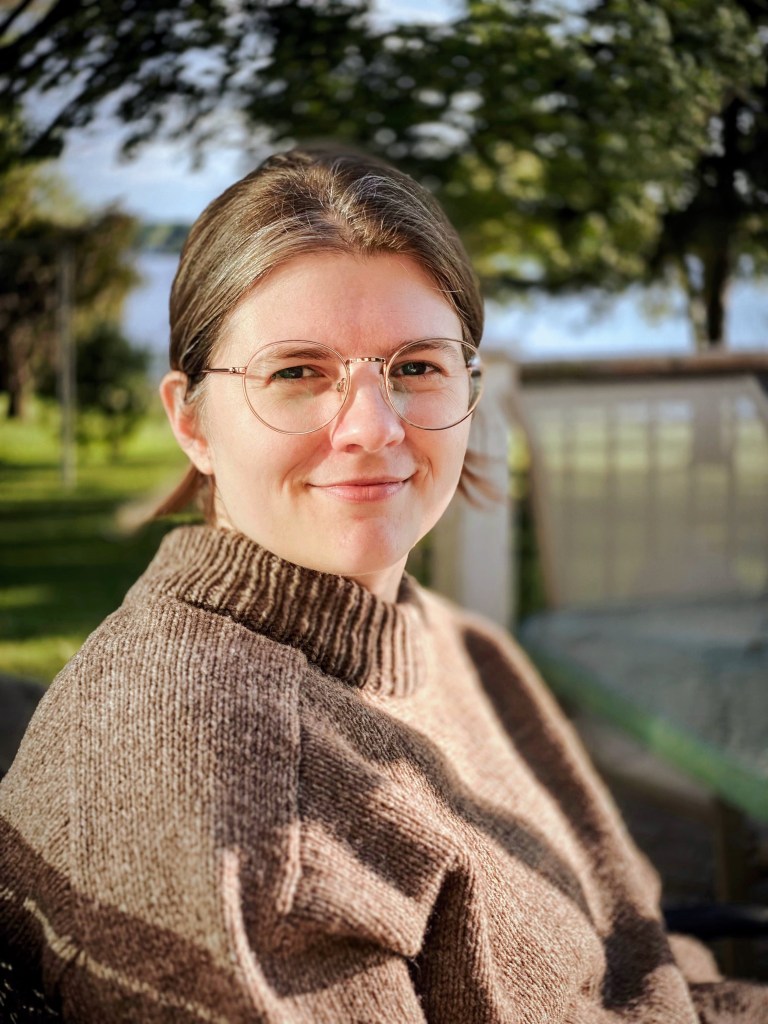 Musicologist, Sarah Koval, a woman with glasses and a brown sweater smiles at the camera, surrounded by greenery and a serene outdoor background.